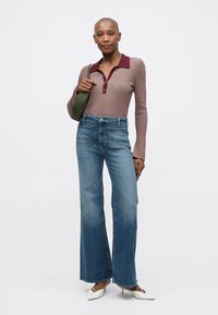 Γυναίκα που φοράει ένα μακρυμάνικο πλεκτό τοπ σε μοβ απόχρωση με μπορντό γιακά, ψηλόμεσα παντελόνια με φαρδιές μανίκες, λευκά τακούνια και κρατάει μια ελαιόχρωμη τσάντα ώμου.