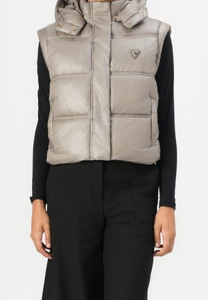 Veste sans manches matelassée taupe avec capuche portée sur un haut noir à manches longues et un pantalon noir, mains détendues le long du corps.
