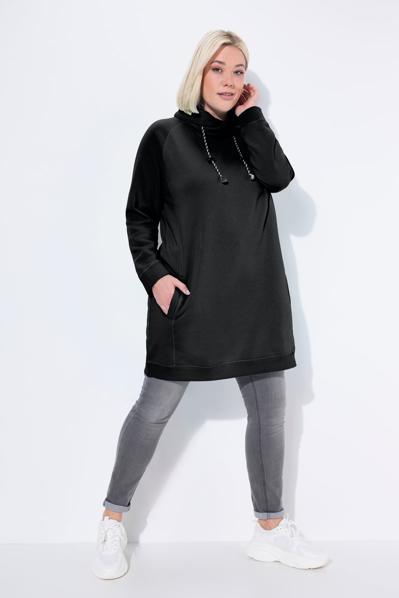 Donna in piedi su sfondo bianco che indossa una felpa nera lunga con coulisse, jeans skinny grigi risvoltati e sneakers bianche.