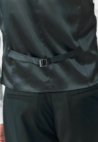 Fekete selyemmel bevont mellény sima textúrával, középen állítható hevederrel, fém csattal. Fekete nadrággal és fehér inggel párosítva.