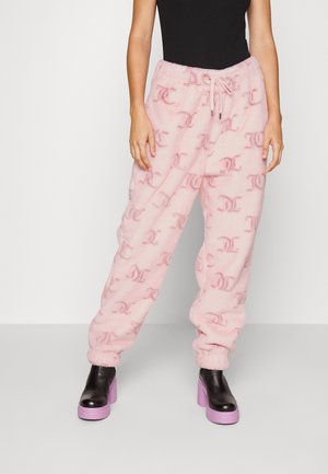 Persoon draagt roze fleecebroek met trekkoord en monogrampatroon, zwarte top en zwarte laarzen met paarse plateauzolen.