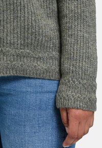 Khakifarbener Strickpullover mit gerippter Textur und dicken Bündchen, kombiniert mit blauen Jeans. Das Material wirkt warm und weich.