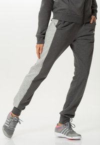 Personne portant un pantalon de jogging gris foncé avec des bandes latérales gris clair et des chaussures de sport grises avec des touches roses, debout avec une jambe croisée devant.