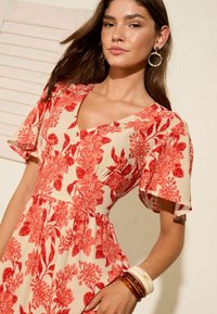 Rochie florală cu un fundal crem, având modele florale roșii, decolteu în V, mâneci scurte și ușoare, și detalii cu nasturi pe bust.