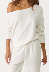 Sweat-shirt blanc à épaules dénudées avec manches longues, fait de matière douce. Associé à un pantalon assorti de couleur claire, avec une coupe décontractée.
