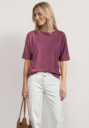 Blondynka w luźnej fioletowej bluzce z krótkim rękawem, białych spodniach z wysokim stanem, uśmiechnięta, trzymająca brązową plecioną torbę przy boku.