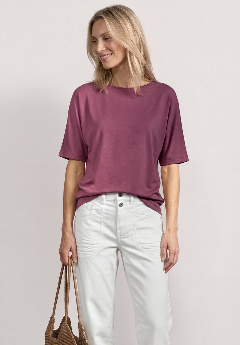 Femme blonde portant un haut violet ample à manches courtes, un pantalon blanc taille haute, souriante et tenant un sac cabas tressé marron à ses côtés.