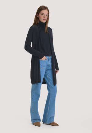 Frau trägt einen schwarzen Rollkragenpullover und langen Cardigan mit hellblauen weit geschnittenen Jeans sowie braunen spitzen Schuhen, steht mit der Hand in der Tasche.