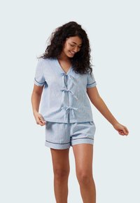 Světle modrý pyžamový set obsahující krátký rukáv a top s V-výstřihem a zavazováním, doplněný o odpovídající šortky, zdobený tmavým lemem a jemným vzorem.