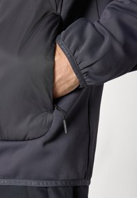 Temno siva jakna z gladko teksturo, ki ima žep z zadrgo in elastične manšete. Oblikovana za udobje in funkcionalnost.