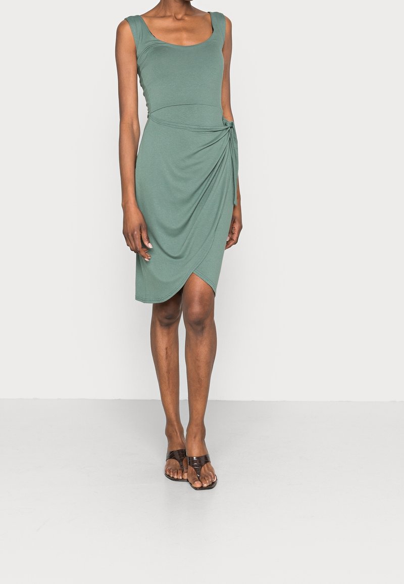 Femme portant une robe portefeuille sans manches vert sauge et des sandales plates marron, se tenant devant un fond clair uni.