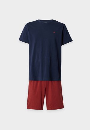 Man standing barefoot against plain background wearing a navy blue t-shirt and red knee-length shorts.