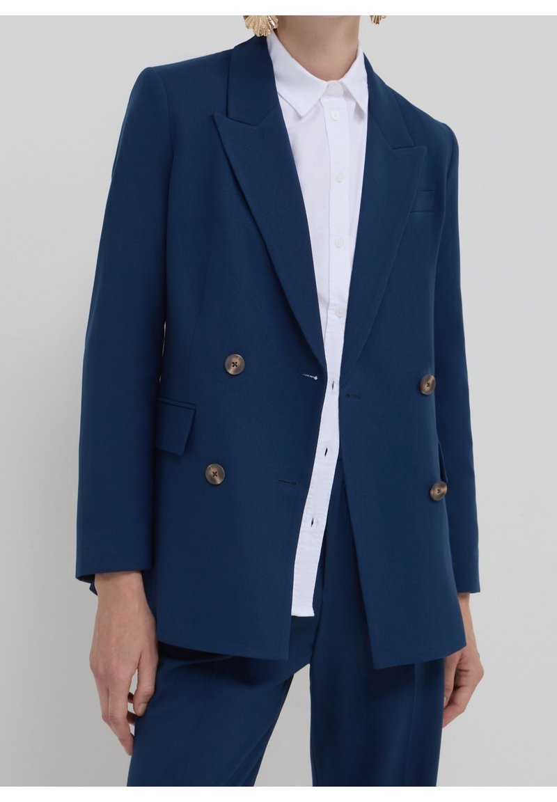 Femme portant un blazer croisé bleu marine à boutons marron sur une chemise blanche boutonnée, assorti à un pantalon bleu marine.