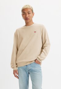 Beige sweatshirt met een ronde halslijn, geribbelde mouwen en zoom, met een klein rood logo op de borst; gecombineerd met lichtblauwe jeans.