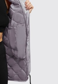 Veste rembourrée violet clair avec un motif matelassé, présentant une poche intérieure. Tissu nylon lisse, fermeture éclair latérale avec tirette de marque.