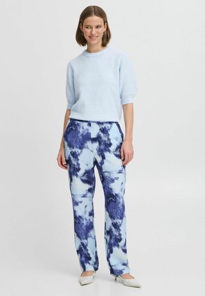 Helles blaues Strickoberteil mit kurzen Ärmeln, kombiniert mit blauen und weißen gemusterten Hosen mit einem aquarellartigen Design; die Absätze sind cremefarben.