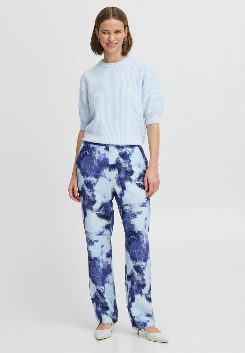 Helles blaues Strickoberteil mit kurzen Ärmeln, kombiniert mit blauen und weißen gemusterten Hosen mit einem aquarellartigen Design; die Absätze sind cremefarben.