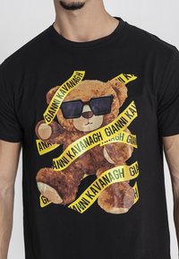 Camiseta negra con un gráfico de un oso de peluche marrón usando gafas de sol, rodeado por un texto en cinta amarilla que dice "GIANNI KAVANAGH."