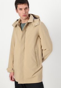 Beige vandtæt jakke med hætte, lynlås foran, sidelommer og justerbare manchetter. Glat tekstur og minimalistisk design.