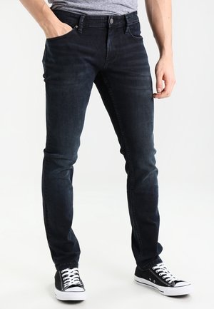 Mand iført sorte slim fit-jeans og sorte lærredssneakers med hvide snørebånd, stående med den ene hånd i lommen.