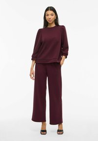 Burgunderfarget genser med pufferm, sammen med vidtrekkende bukser. Myk tekstur, uformelt design og rund hals. Svart sandaler med åpen tå.