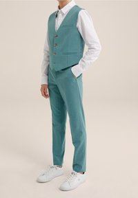 Tyrkysová vesta se třemi knoflíky, přední kapsou, v kombinaci s ladícími kalhotami a bílými teniskami. Hladká látka, přiléhavý střih.