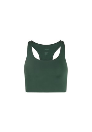 Vörös hajú nő áll, karjait felemelve, sötétzöld sportmelltartót és magas derekú kerékpáros rövidnadrágot visel, fehér zoknival és fehér tornacipővel, egyszínű háttér előtt.