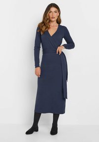 Marineblaues Wickelkleid mit langen Ärmeln, V-Ausschnitt und Taillengürtel. Glatte Stoffqualität, knielang, kombiniert mit schwarzen Knöchelstiefeln.