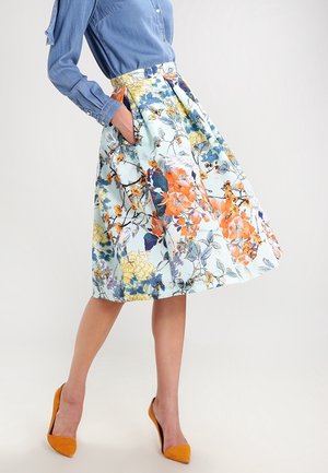 Woman wearing a pleated, knee-length floral skirt with pockets, paired with a blue long-sleeve shirt and mustard-yellow high heels.