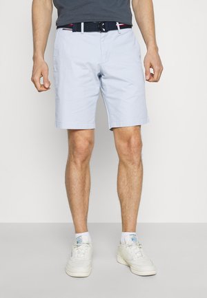 Homme portant un short bleu clair, une ceinture marine, une chemise noire, des chaussettes blanches et des baskets blanches, debout devant un fond uni.