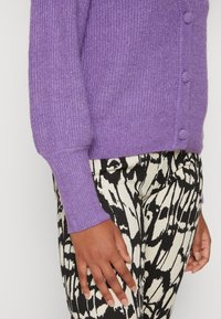 Lila stickad cardigan med ribbade manschetter och knappar, kombinerad med svarta och vita mönstrade byxor som visar en texturerad väv.