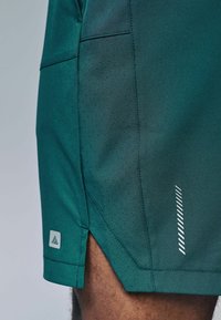 Grønne atletiske shorts lavet af åndbart stof, med en perforeret sidepanel, skråt undermal og hvide refleksdetaljer.