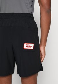 Shorts de sport noirs avec une taille élastique, dotés d'une poche plaquée ornée du logo "Nike" en rouge et blanc. Tissu à la texture lisse.