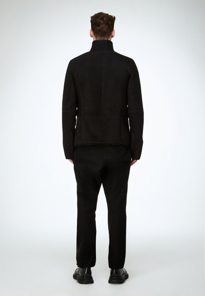 Schwarze Wildlederjacke mit hohem Kragen, klaren Linien und Nahtdetails, kombiniert mit schwarzer Hose und Schuhen, von hinten gezeigt.