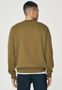 Sudadera marrón de cuello redondo con mangas largas, puños y dobladillo acanalados, llevada sobre una camisa blanca, combinada con jeans azul oscuro.