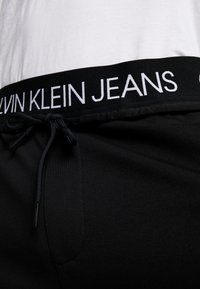 Svarta joggingsbyxor med dragsko i midjan, med en svart midjeband som har vit "CALVIN KLEIN JEANS" branding. Mjuk, elastisk material.
