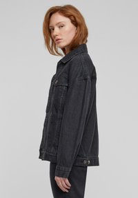 Fiatal vörös hajú nő, aki túlméretezett fekete farmer dzsekit és fekete nadrágot visel, oldalra fordulva, egyszerű háttér előtt áll.