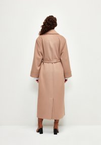 Abrigo largo con cinturón en suave tejido beige, con una abertura en la parte posterior, que presenta mangas anchas y un cuello clásico. Textura suave y minimalismo en los detalles.