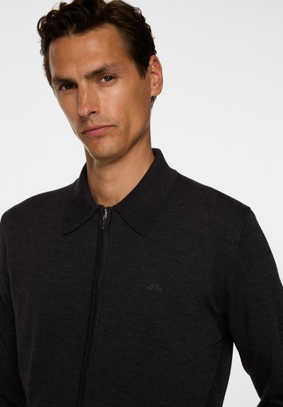Uomo con capelli scuri che indossa un maglione nero con zip e colletto su uno sfondo bianco semplice.