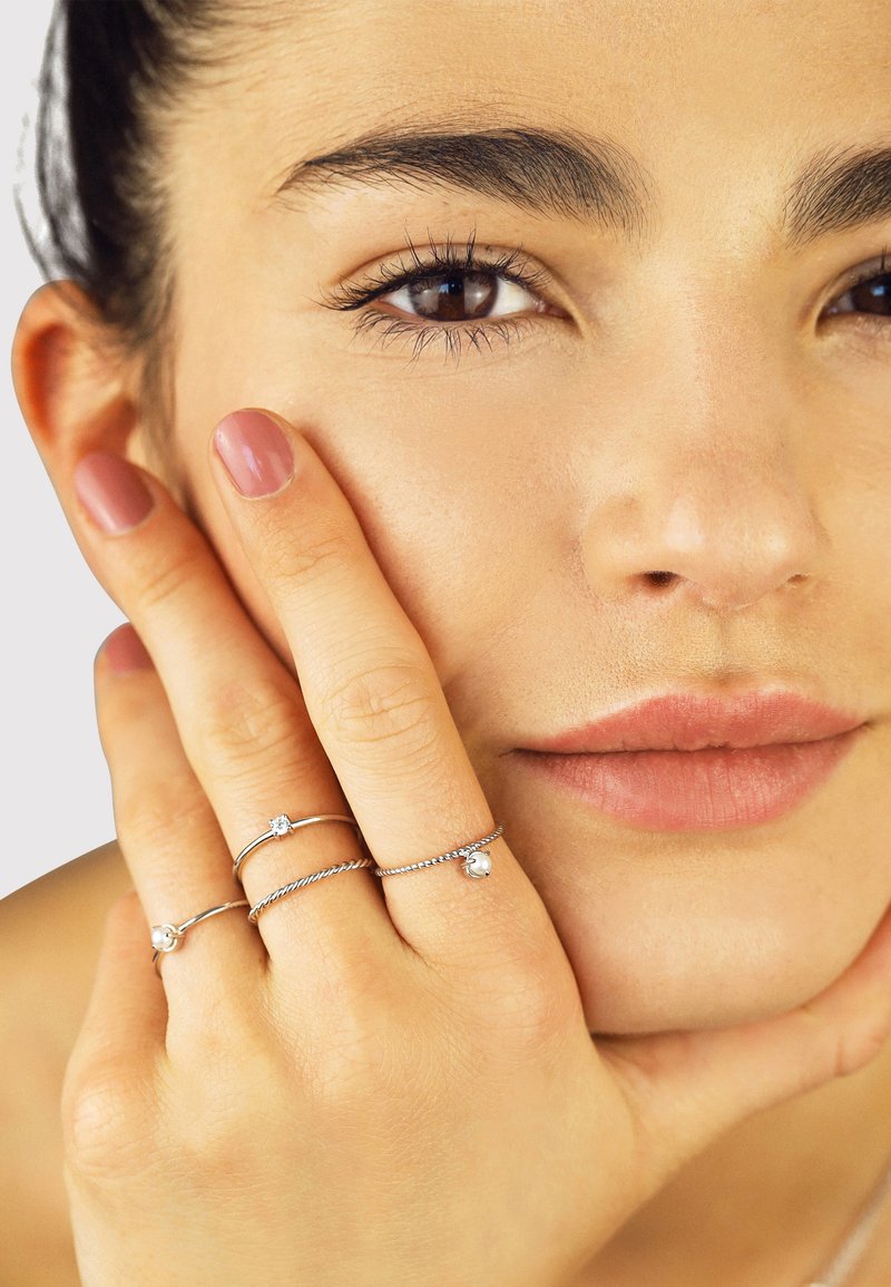 Drei silberne Ringe mit verschiedenen Designs, darunter ein gedrehter Ring und Akzentsteine, getragen an einer Hand, die nahe einem Gesicht mit natürlichem Make-up ruht.
