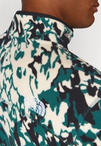 Mand iført højhalset fleece med grønne, sorte og hvide abstrakte mønstre samt hvidt broderet "The North Face" logo på skulderen.