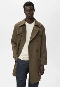 Bruine double-breasted trenchcoat van een gladde stof, met een brede kraag, grote knopen en een losse pasvorm, gedragen over een licht shirt.