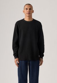 Black long-sleeve ribbed knit top with a crew neckline, relaxed fit, and textured surface, paired with dark blue wide-leg jeans.