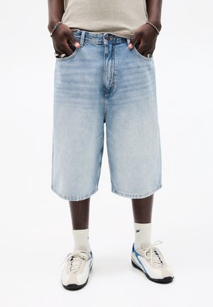 Persona che indossa pantaloncini lunghi in denim azzurro chiaro, top beige, calzini bianchi e scarpe da ginnastica bianche con dettagli blu, in piedi con le mani in tasca.