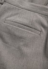 Pelēka auduma kabatas detaļa ar smalku tekstūru un tīrām šuvēm, kurā ir viena veldēta kabata, novietota aizmugurē.