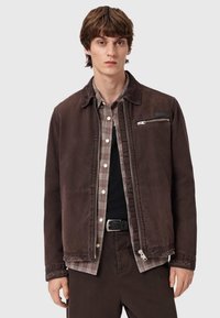 Brązowa kurtka z kołnierzem ze sztruksu, zapinana na zamek kieszeń oraz subtelna, postarzona tekstura, nałożona na koszulę w kratę i czarną koszulkę.