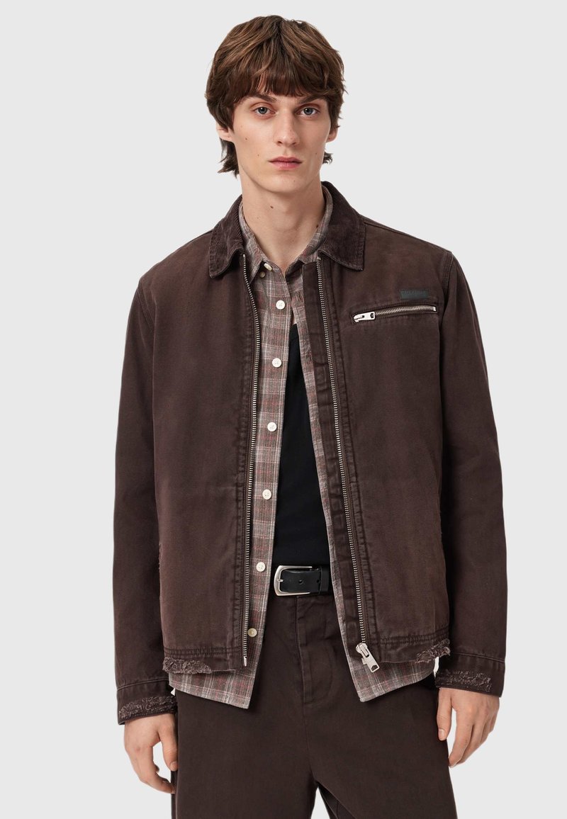 Brązowa kurtka z kołnierzem ze sztruksu, zapinana na zamek kieszeń oraz subtelna, postarzona tekstura, nałożona na koszulę w kratę i czarną koszulkę.