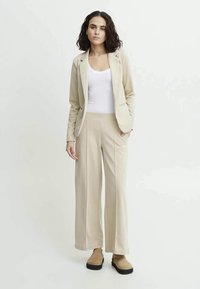Woman with dark hair wearing beige blazer and wide-leg trousers over white top, standing with one hand in pocket on white background.