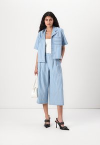Blazer cropped a righe blu chiaro con maniche corte, abbinato a pantaloni larghi coordinati. La modella tiene una borsa bianca e indossa tacchi neri.