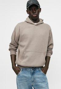 Ανοιχτό καφέ φούτερ με μπροστινή τσέπη, ραμμένα μανίκια και κουκούλα με κορδόνι. Συνδυάζεται με ανοιχτό μπλε τζιν παντελόνι. Απλός σχεδιασμός.